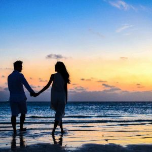 Work With Me A romantic couple walks hand in hand on a tropical beach at sunset, enjoying a serene moment together.